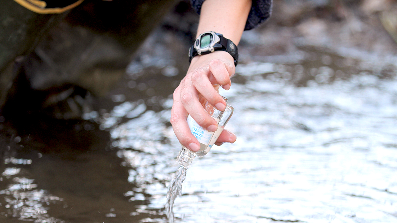 Hand reaching over river holding a glass collecting do water samples.
